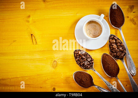 Verschiedene Kaffeebohnen und Begründung Anzeige in Löffel aus der unteren rechten Ecke mit Espresso in der Tasse auf einem gelben Holz Hintergrund mit vignette und Stockfoto