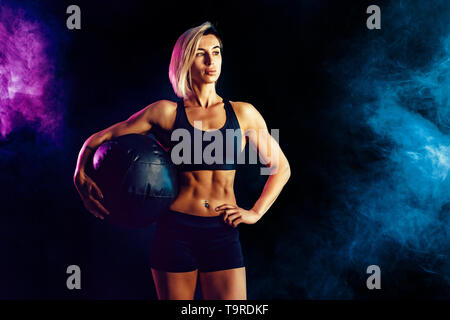 Sportliche Blondine in modischer Sportswear mit Medizinball posieren. Foto von muskulöse Frau auf dunklem Hintergrund mit Rauch. Kraft und Motivation. Stockfoto