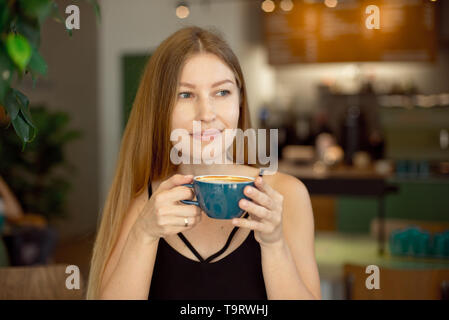 Junge schöne Blondine mit langen Haaren in einem Charme Top und Jeans Getränke Kaffee und schaut aus dem Fenster Stockfoto