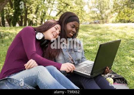 Paar der multi-ethnischen Mädchen Schüler sitzen auf der Wiese am Notebook Stockfoto