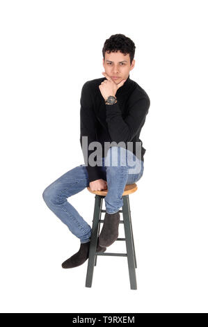 Schöne junge Mann in einem schwarzen Pullover und Jeans sitzt auf einem Bar Stuhl mit einer Hand unter sein Kinn, auf weißem Hintergrund Stockfoto