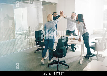 Vielfältige Gruppe von lächelnden Arbeit Kollegen um ein Büro Tisch und hohe FIVING während einer Sitzung ansehen Stockfoto