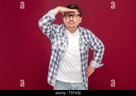 Portrait von aufmerksamen gut aussehender Mann mittleren Alters in lässig-kariertem Hemd und Brille stehend mit der Hand an der Stirn und auf der Suche nach Etwas. indo Stockfoto