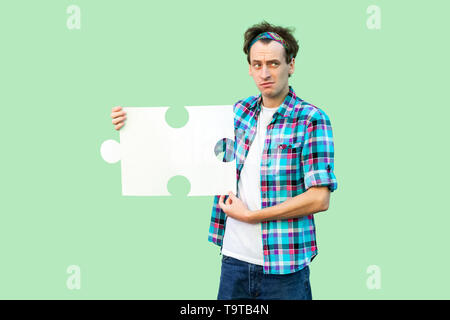 Portrait von Traurigkeit junger Mann in kariertem Hemd und mit großen Stück des Puzzles, denken über die Lösung seines Problems. Hallenbad, isoliert, Stockfoto