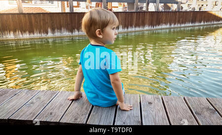 Adorable 3 Jahre altes Kleinkind Junge sitzt am Ufer in kleinen europäischen Stadt an der sonnigen Sommertag Stockfoto