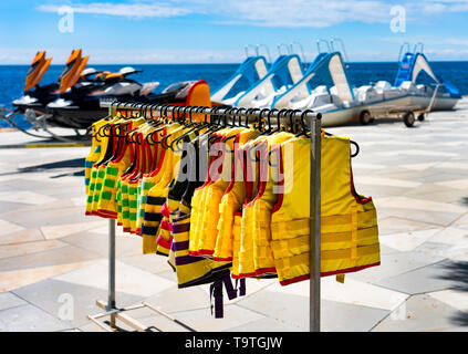 Eine Menge Schwimmwesten hängen am Strand. Wasser, Scooter und Tretboot im Hintergrund. Das Konzept der Sicherheit. Stockfoto