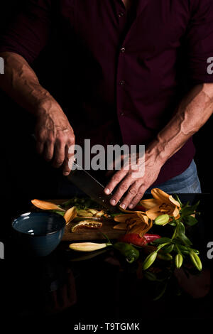 Mann schneidet Lilien und farbige Paprika auf Holz Schneidebrett Stockfoto