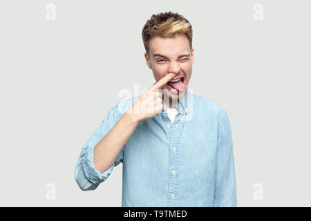 Portrait von Crazy hübscher junger Mann in Hellblau shirt stehend, seine Nase bohren, winking, Zunge raus, und wenn man die Kamera mit lustigen Gesicht. die Pflege von Stockfoto