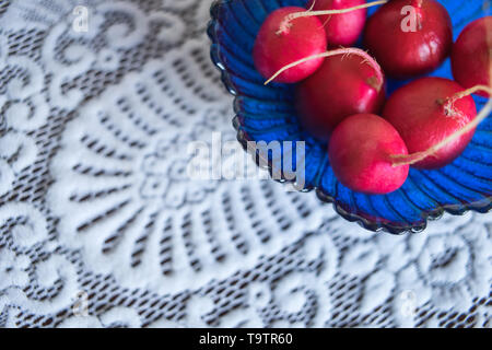 Frische reife Radieschen in einem blauen Glas salat Schüssel auf eine weiße Tischdecke mit einem zarten Muster. Platz kopieren Stockfoto