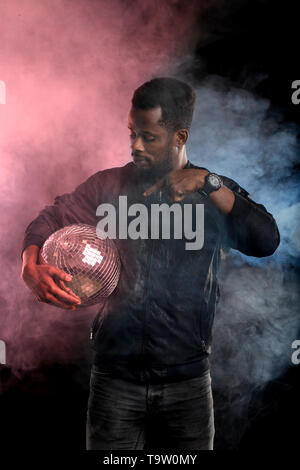 Junge coole schwarze Mann mit Disco Kugel über schwarzen Hintergrund. Vertikale Portrait von african american Kerl durch die Rosa und blauen Rauch im Dunkeln. Unterhaltung Stockfoto