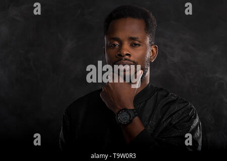 Headshot von schweren Dunkelhäutige männliche Holding Kinn geheimnisvoll Kamera suchen, in schwarz windjacke Jacke, wristwatces gekleidet, gegen die ständigen Stockfoto