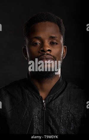 Sehr detaillierte Closeup Portrait von attraktiven jungen afrikanischen Mann, schwarze Jacke mit Kamera mit ernsten und nachdenklichen Ausdruck auf dunklen Stockfoto