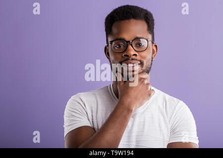 Fröhlich afrikanische Kerl in Gläser im weißen T-Shirt lächelnd in gute Laune berühren Kinn, während im Studio über violette Hintergrund posiert gekleidet. Gesichtsbehandlung Stockfoto