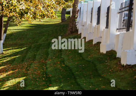 Herbst Hintergrund. Herbst Garten mit Laub auf dem grünen Rasen und einem weißen Zaun an einem sonnigen Tag. Stockfoto