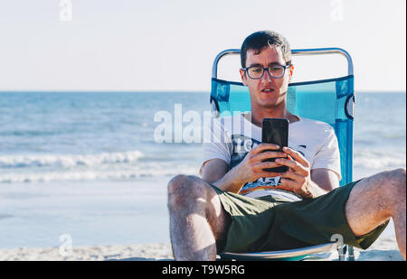 Porträt der glückliche junge Mann in der Hängematte mit Meer Blick auf Smartphone am Strand zu sitzen mit dem Meer im Hintergrund Stockfoto