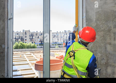 Ein Arbeiter in einem orange Helm installiert eine doppelt verglaste Fenster, in einem neuen Haus, vor dem Hintergrund der Konstruktion. Stockfoto