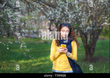 Junge smilling stehende Frau in einem blühenden Garten und schreibt auf Handy. Kirschblüten. Porträt der schönen Frau Stockfoto