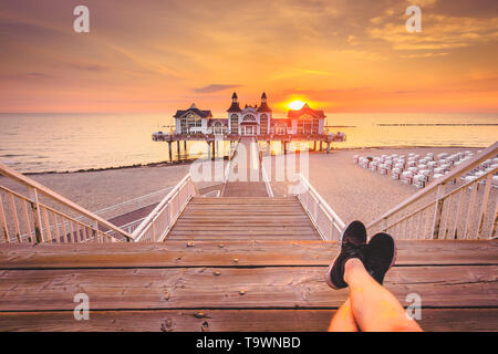 Junge Mann sitzt auf hölzernen Pier genießen Sie einen wunderschönen Sonnenaufgang am berühmten Seebrucke Sellin in Ostseebad Sellin, Insel Rügen, Ostsee, Deutschland Stockfoto