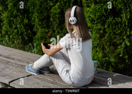 Frau Hören von Musik über Kopfhörer und mit einem Smartphone. Eine Frau sitzt auf der Bank im Park an einem sonnigen Frühlingstag. Modische Frauen Stockfoto