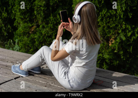 Junge Mädchen, das Hören von Musik über Kopfhörer und Suchen in einem Smartphone. Eine Frau sitzt auf der Bank im Park an einem sonnigen Frühlingstag Stockfoto