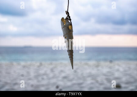 Dieses einzigartige Foto zeigt eine Muschel hängend an einer Schnur und im Hintergrund können Sie den Strand und den Sonnenuntergang auf den Malediven sehen Stockfoto