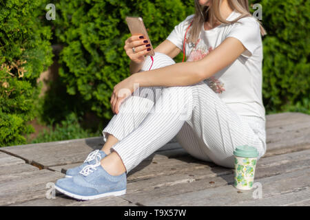 Mädchen suchen in Smartphone und auf der Bank sitzen neben einer Tasse Kaffee im Park an einem sonnigen Frühlingstag. Modische Frauen in gestreiften Hosen Stockfoto