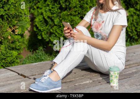 Mädchen mit einem Smartphone. Eine Frau auf der Bank sitzen neben einer Tasse Kaffee im Park an einem sonnigen Frühlingstag. Modische Frauen in gestreiften Hosen Stockfoto