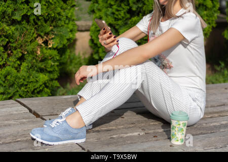 Frau Arbeit auf einem Smartphone und auf der Bank sitzen neben einer Tasse Kaffee im Park an einem sonnigen Frühlingstag. Modische Frauen in gestreiften Hosen Stockfoto