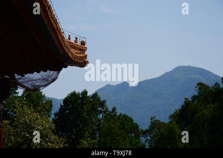 Traditionelle orange kunstvollen chinesischen Fachwerkbalken Blick auf BEWALDETE asiatischen Berge. Grüne Bäume im Vordergrund, Hitzeflimmern und blauer Himmel. Figuren geschnitzt auf oder Stockfoto