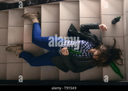 Bewusstlose junge Frau mit leeren Flaschen liegend auf Treppen. Konzept des Alkoholismus Stockfoto