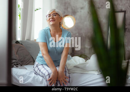Lächelnde Frau tragen Brillen und Pyjama sitzen auf dem Bett Stockfoto