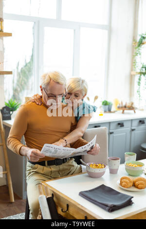 Liebevolle Frau zärtlich umarmt einen Ehepartner beim Zeitung lesen Stockfoto