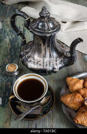 Schwarzen Kaffee in vintage Schale, Croissants auf einem alten Weißblech und Antik Silber Kaffee Topf auf einem alten, dunklen Holzbrettern Stockfoto