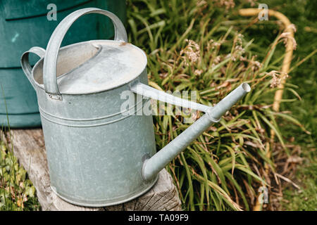 Alte Metall Gießkanne auf einem Verwitterten hölzernen Bar vor einem Wasserbehälter in einer grünen Frühling Garten Stockfoto