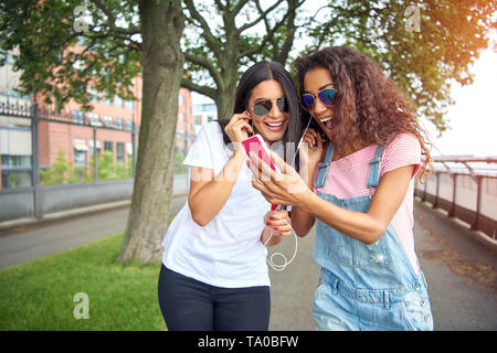 Zwei attraktive junge Freundinnen lächelnd und zusammen gehen hinunter eine Promenade im Sommer Hören von Musik über Kopfhörer Stockfoto