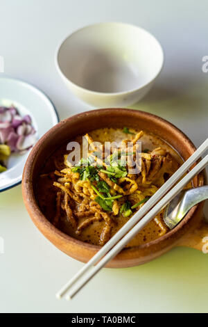 Die nordinsel thailändisches Gericht khoa Soi mit Spiegelei Nudeln und Stäbchen in einen Tontopf und einige Garnieren im Hintergrund, Thailand Stockfoto
