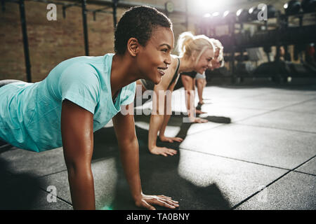 Lächelnde junge afrikanische amerikanische Frau in Sportkleidung pushups tun während eines Trainings Klasse auf dem Boden einer Turnhalle Stockfoto