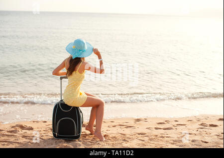 Reisen Urlaub und Personen Konzept - Frau Tourist in der Nähe von dem Meer sitzen auf Koffer und in Hut und beobachten die Sonne und alle Daumen hoch Stockfoto