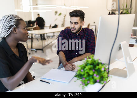 Junge schöne afrikanische Frau mit einem Lächeln mit ihrer indischen Kollegen diskutieren, während im Büro stehen Stockfoto