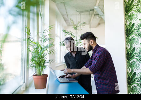 Junge schöne afrikanische Frau pointing at laptop mit einem Lächeln und etwas diskutieren mit ihren indischen Kollegen, während im Büro stehen Stockfoto