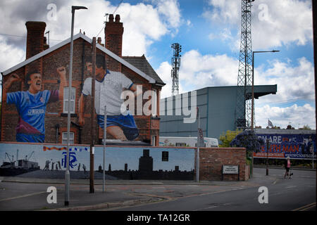 Eine Frau, die zu Fuß einen Hund hinter einem Wandbild durch Künstler Paul Curtis auf die giebelseite eines Hauses in Prenton, Wirral mit zwei Tranmere Rovers Spieler. Das Wandbild ist nur außerhalb des Stadion des Clubs, Prenton Park entfernt. Tranmere Rovers gewonnen Förderung zu EFL League 2 aus der Nationalen Liga über einen Play-off in der Saison 2017-8 und kehrte 12 Monate später, am 25. Mai zum Wembley Newport County für einen Platz in der EFL-Liga eine zu Gesicht. Stockfoto