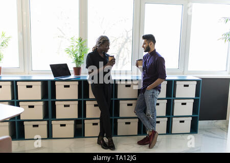 Zwei Unternehmen Mitarbeiter in komfortablen Office während der Kaffeepause mitgeteilt. Schöne indische Kerl im Gespräch mit afrikanischen Frau mit heißen Getränken. Stockfoto