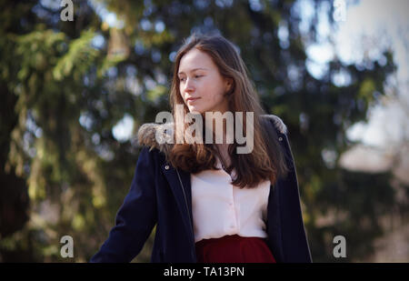 Eine schöne lange blonde Dame in einem Mantel und Rock steht elegant gegen den Wald Hintergrund und schaut etwas mit einem Lächeln. Stockfoto