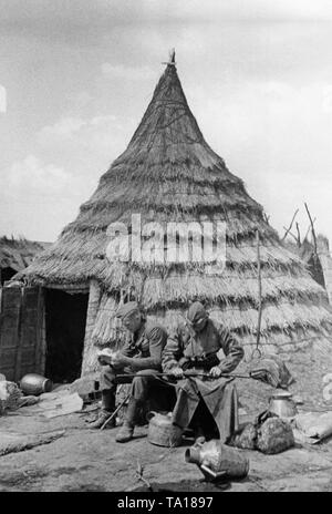 Undatiertes Foto von zwei deutschen Soldaten der Legion Condor, die in einem Waffenstillstand vor eine Strohhütte ruhen. Der Soldat auf der linken Seite ist ein Buch zu lesen, die auf der rechten Seite ist die Reinigung seiner Waffe. Stockfoto