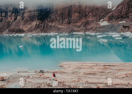 Ein Mann sitzt auf der vorgelagerten Felsen des Oberen Grinnell See und Grinnell Glacier mit niedrigen Festlegung Wolken im Glacier National Park, Montana, USA Stockfoto