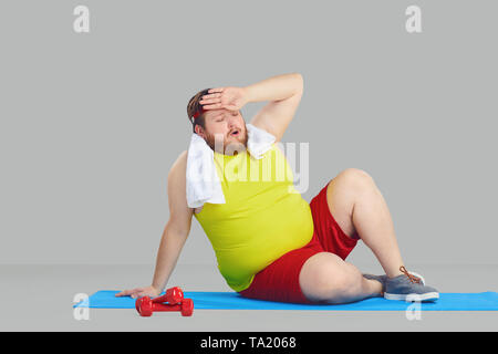 Ein fetter Mann in Sportkleidung müde wischt sich den Schweiß von seiner Stirn auf dem Boden sitzend vor grauem Hintergrund. Stockfoto