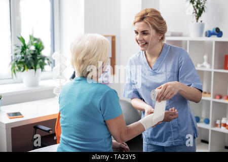 Freudige positive Krankenschwester Verpackung Verband um ihren Patienten hand Stockfoto