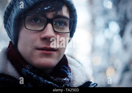 Der Kerl mit Brille und Winterkleidung friert in der kalten Jahreszeit. Schal, Hut und Brille in Schneeflocken Stockfoto