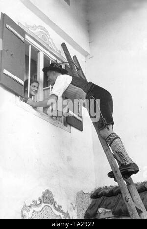 Ein Mann in traditioneller Tracht und Leder hosen Klettert eine Leiter auf dem Fenster der Zimmer seiner Freundin. Stockfoto