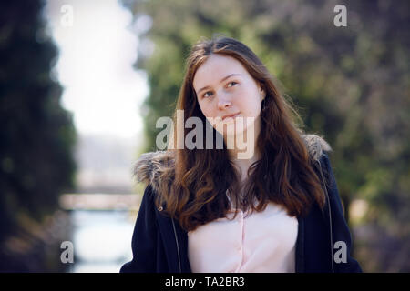 Porträt einer wunderschönen langhaarigen Mädchen in einem Mantel und rosa Bluse steht auf einem sonnigen Tag gegen den Fluss und den Wald. Stockfoto